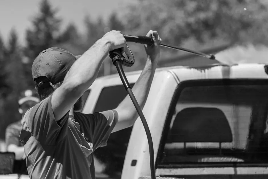 Black and white image of a man using a power spray on a truck outdoors.