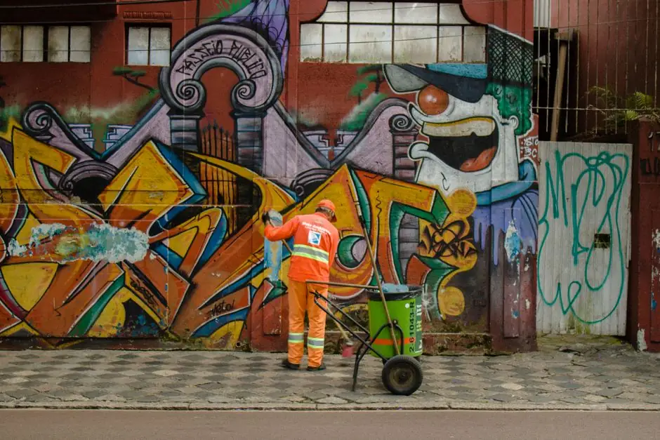 Urban street cleaner in orange uniform working against a vibrant graffiti wall.